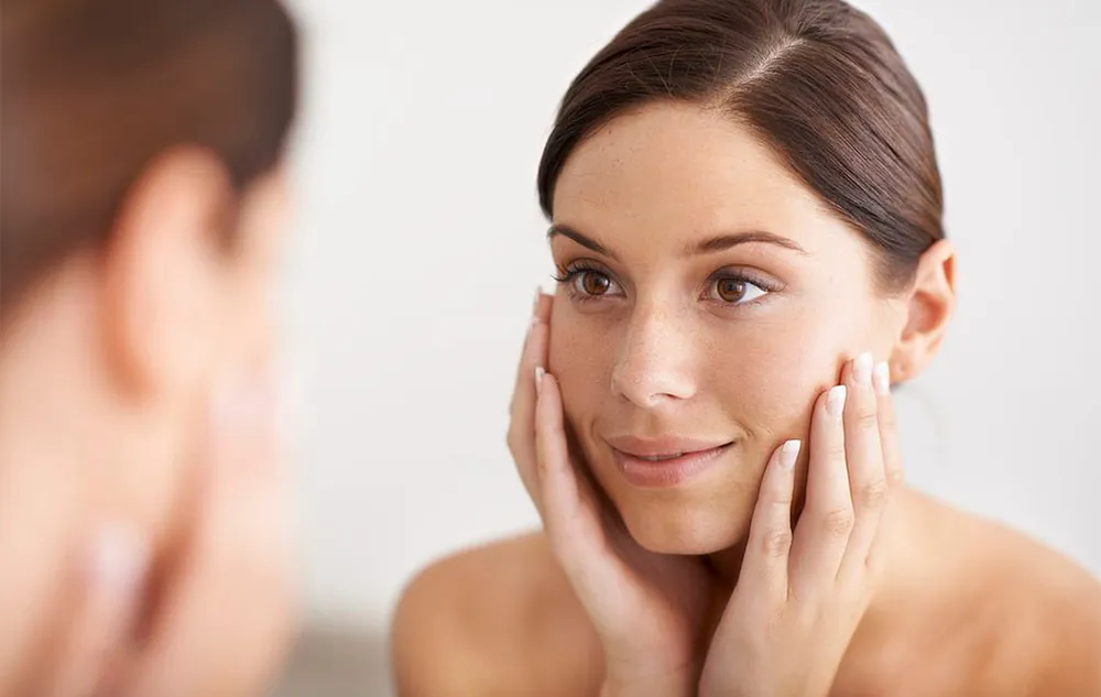 mujer mirándose al espejo con una expresión de satisfacción y con las manos en las mejillas