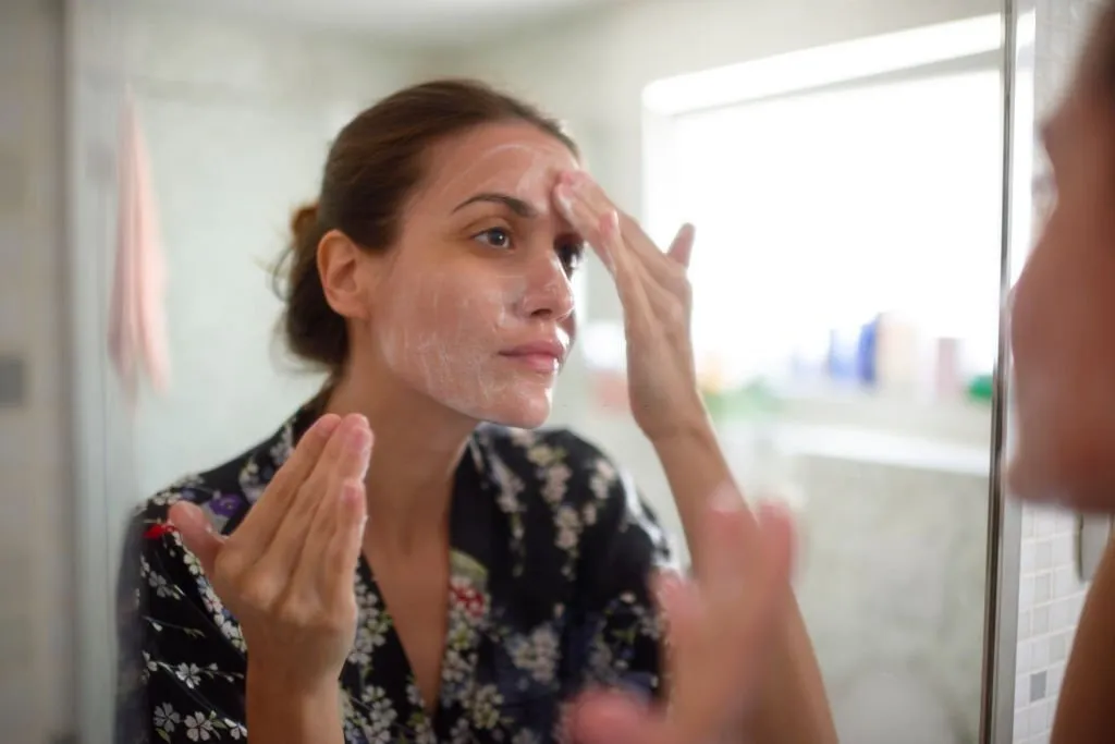 mujer frente a un espejo untándose crema en la cara para cuidar su piel