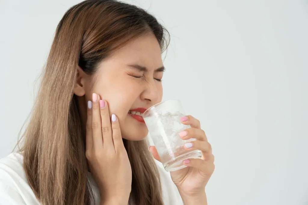 mujer asiática con mano en la mejilla, aquejándose de dolor dental por beber agua fría